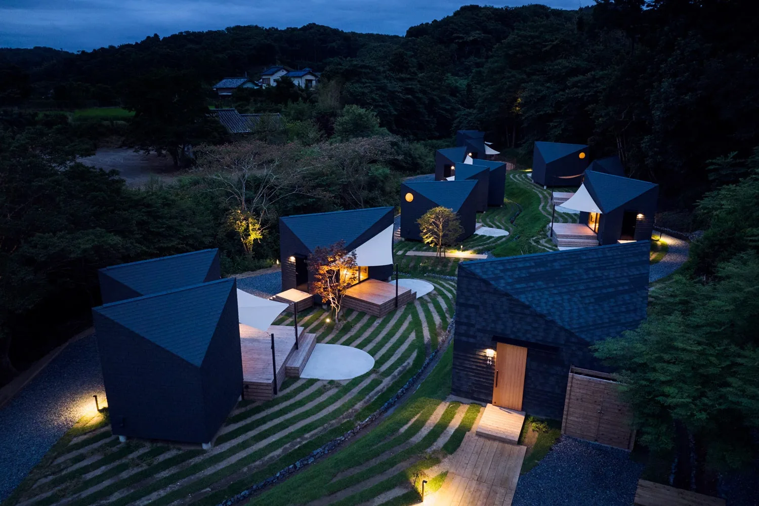 Dark angular cabins lit at dusk on a terraced, grassy hillside