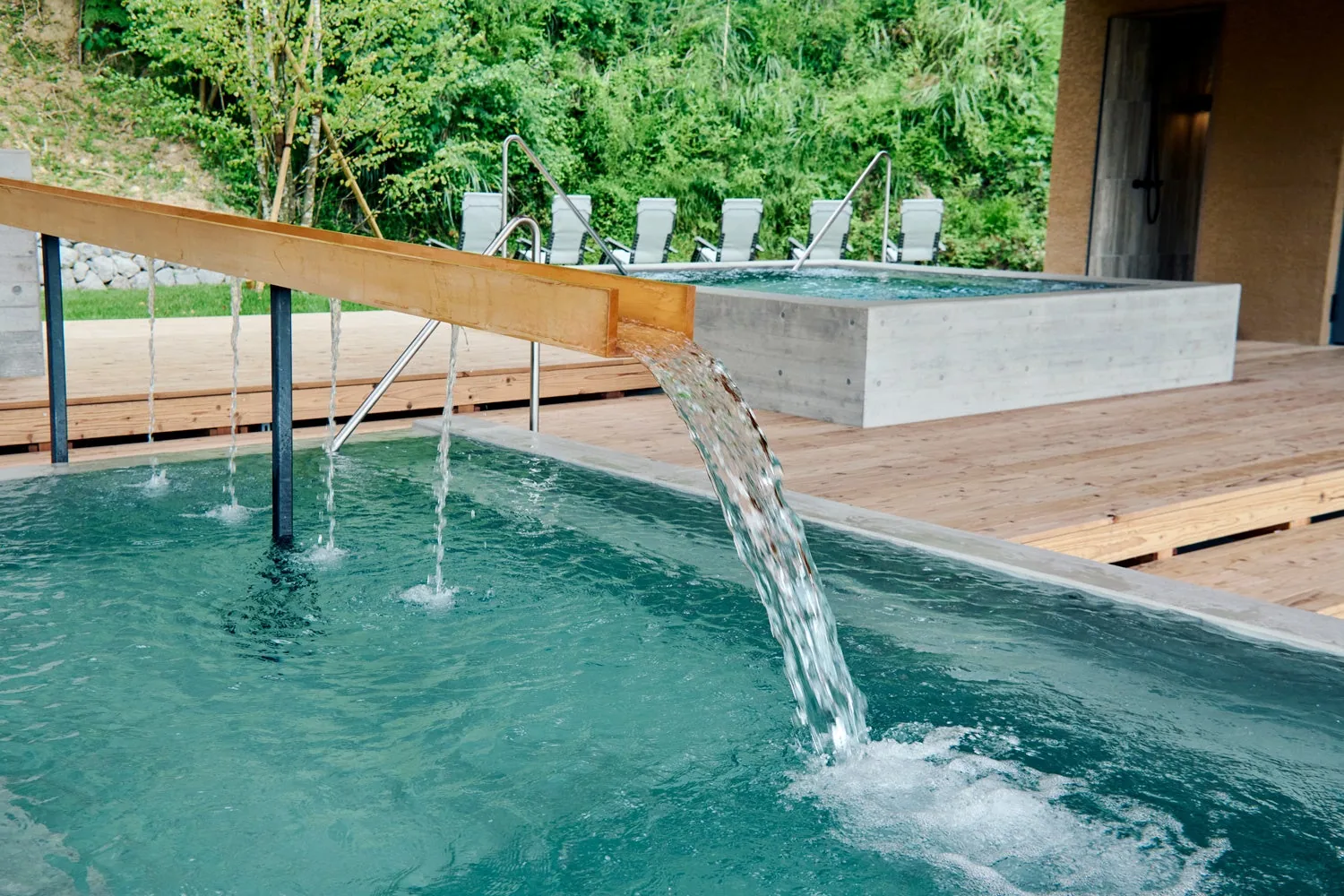 Water pouring from a wooden spout into a blue-green outdoor cold bath, with deck chairs and a second bath behind it