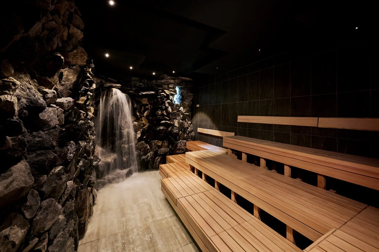 Spring water falling down a dark natural-stone wall beside tiered wooden sauna benches