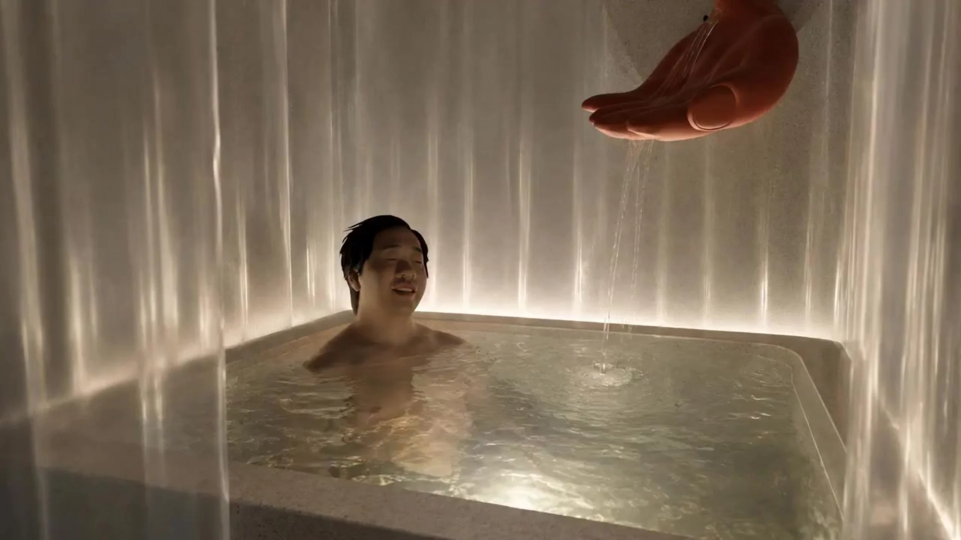 A guest sits in a curtained cold bath beneath water flowing from a large sculpted hand.