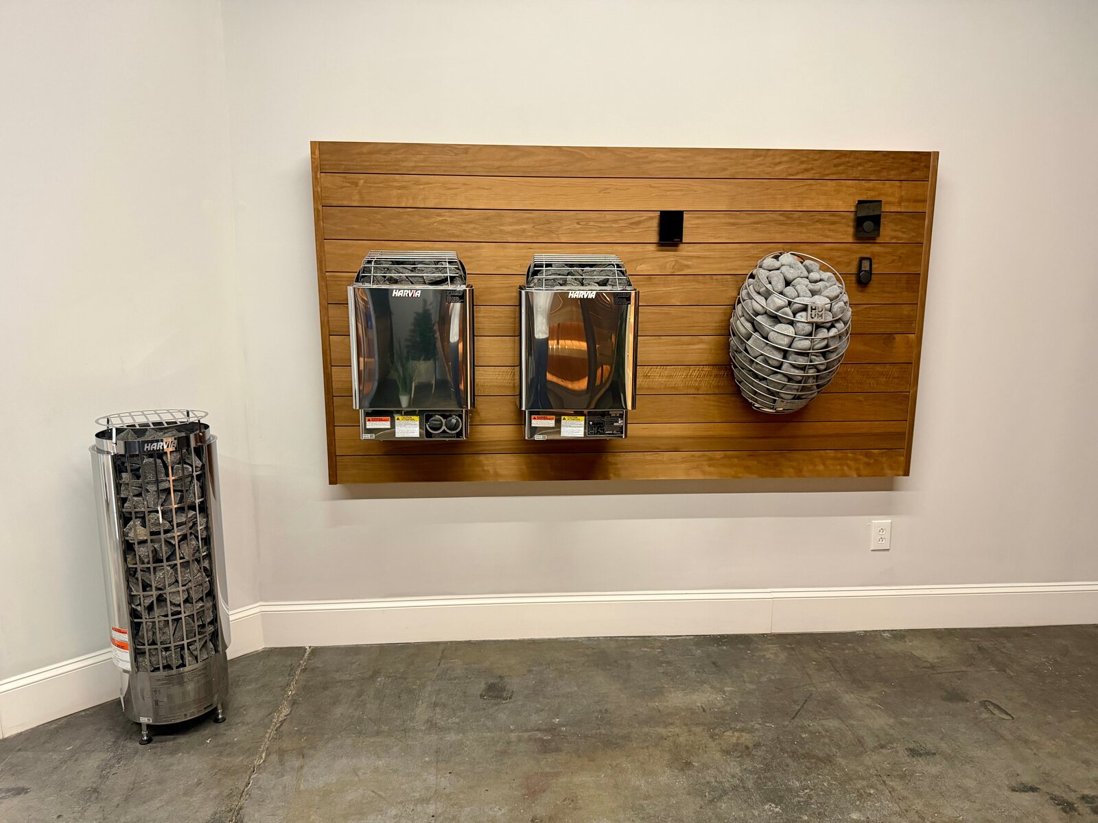 A slatted wood display panel holding two stainless Harvia wall heaters, a round HUUM Hive heater wrapped in a cage of stones and three wall controls, with a tall Harvia Cilindro heater standing on the floor beside it