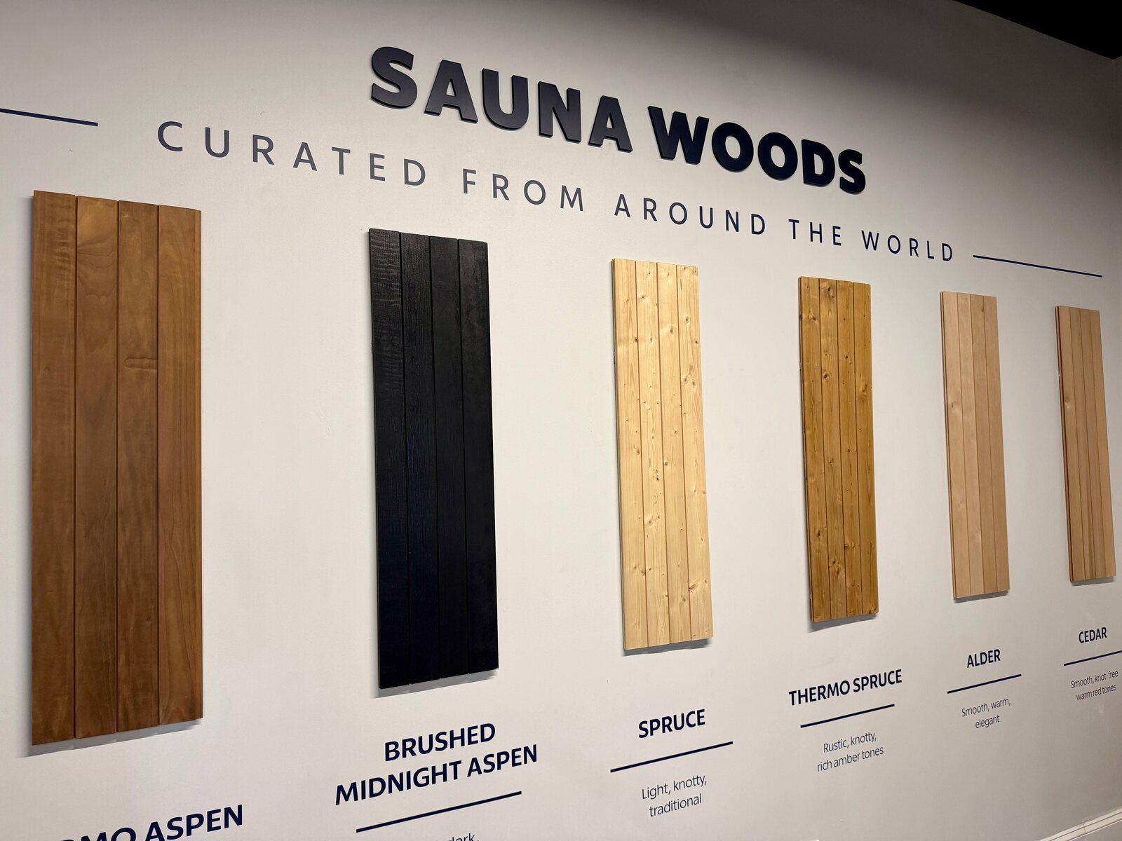 Six vertical sample boards of sauna paneling mounted on a white wall under the words Sauna Woods, Curated From Around the World, from dark brown thermo aspen and black brushed aspen to pale spruce, golden thermo spruce, alder and cedar