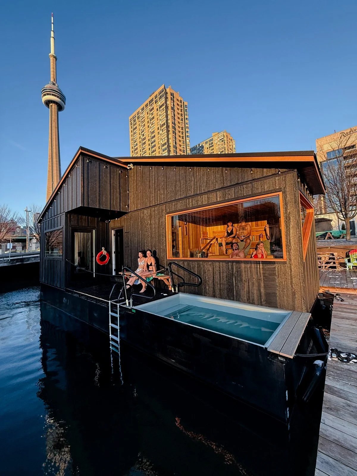 Bathers in a floating sauna and on its deck beside a plunge tub, with Toronto's CN Tower behind it