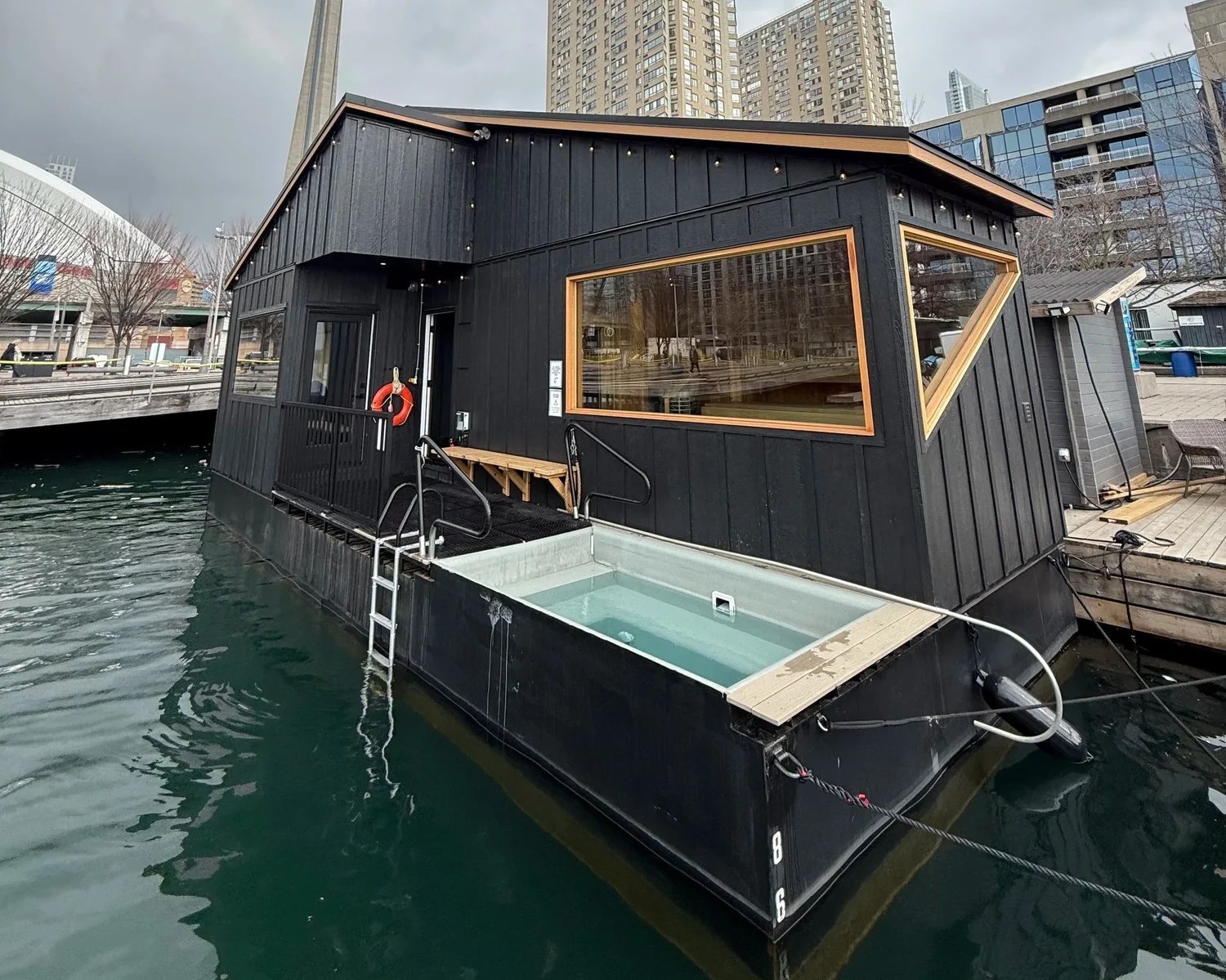 Black-clad floating sauna and rectangular plunge tub moored beside Toronto’s harbourfront