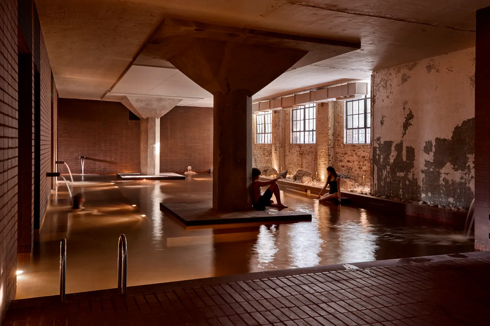 Bathers sit on a platform around a concrete column in a large, warmly lit mineral pool, with original warehouse windows and weathered brick walls behind