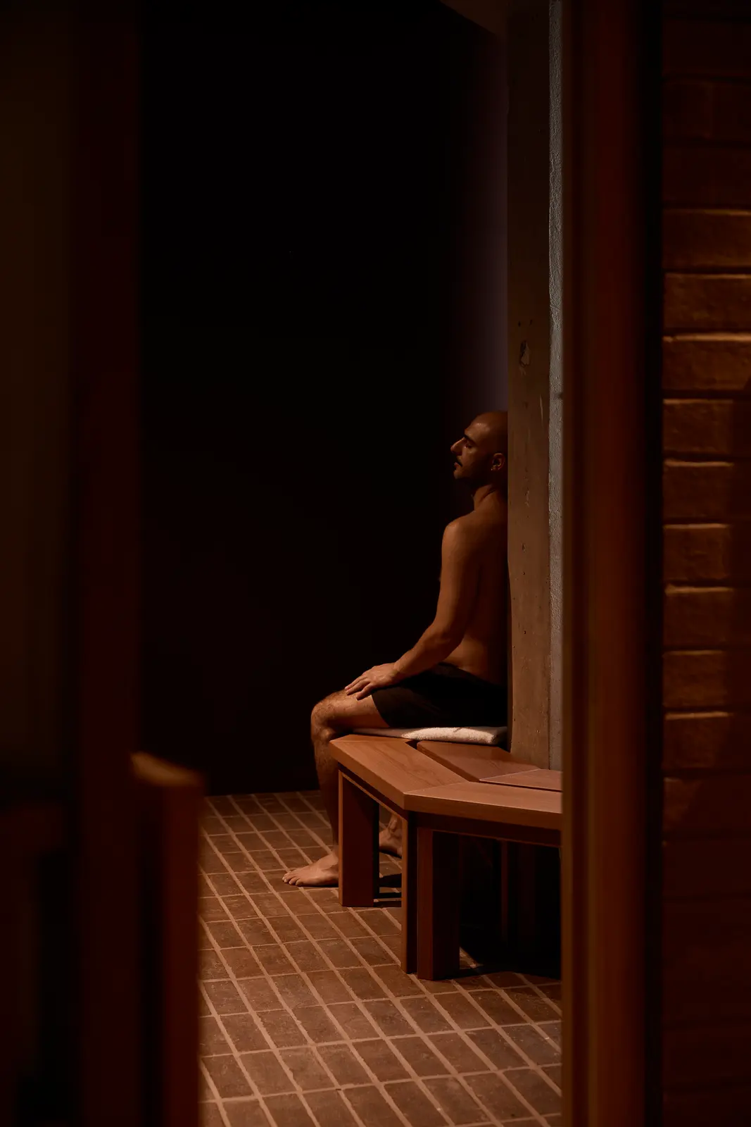 A man in swim shorts sits on a wooden bench against a concrete pillar in a dark room with a brick tile floor, eyes closed