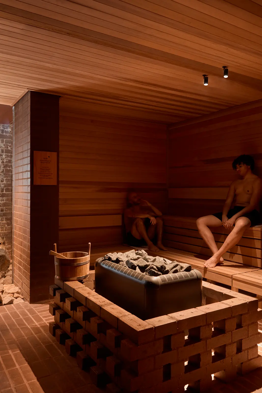 Two bathers sit on wooden benches in a small sauna above a heater filled with grey stones inside a brick enclosure, with a wooden bucket on the brick ledge