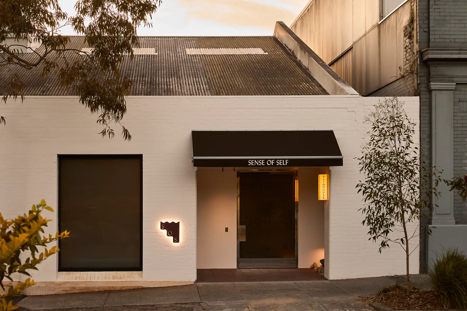 A white-painted brick warehouse with a black awning reading Sense Of Self over the door, a lit SOS wall sign and a native tree out front