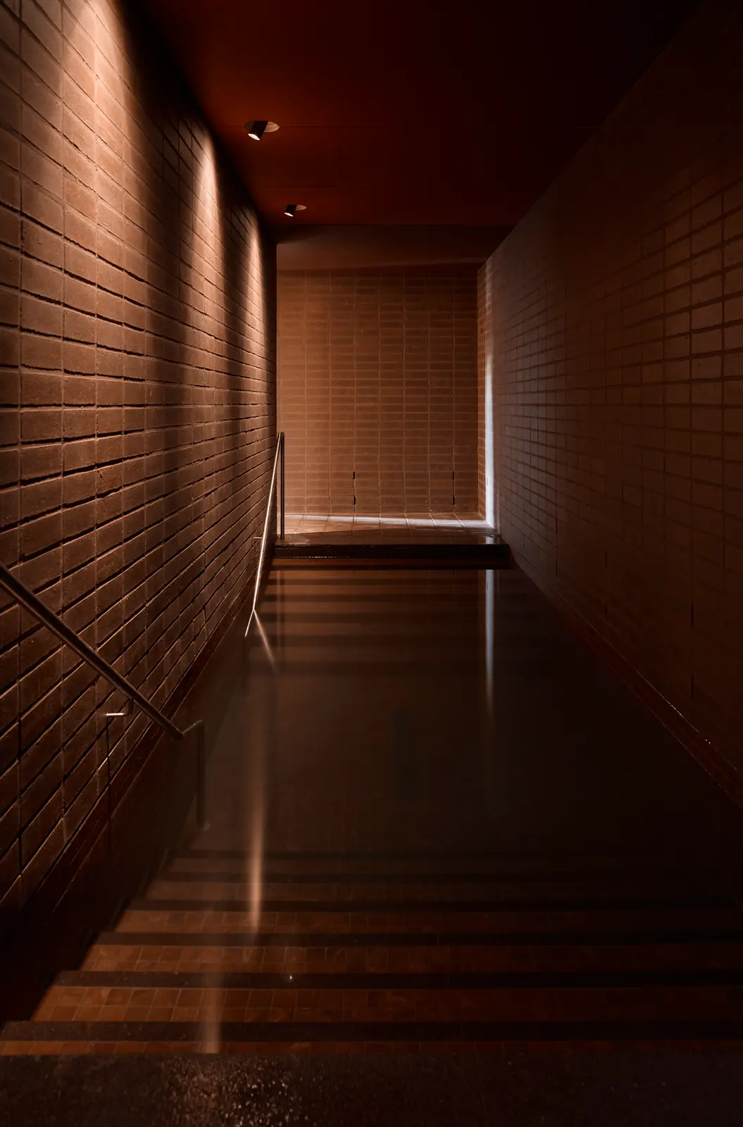 A long, narrow cold plunge channel between dark brick walls, with steps leading down into the water and a handrail along one side