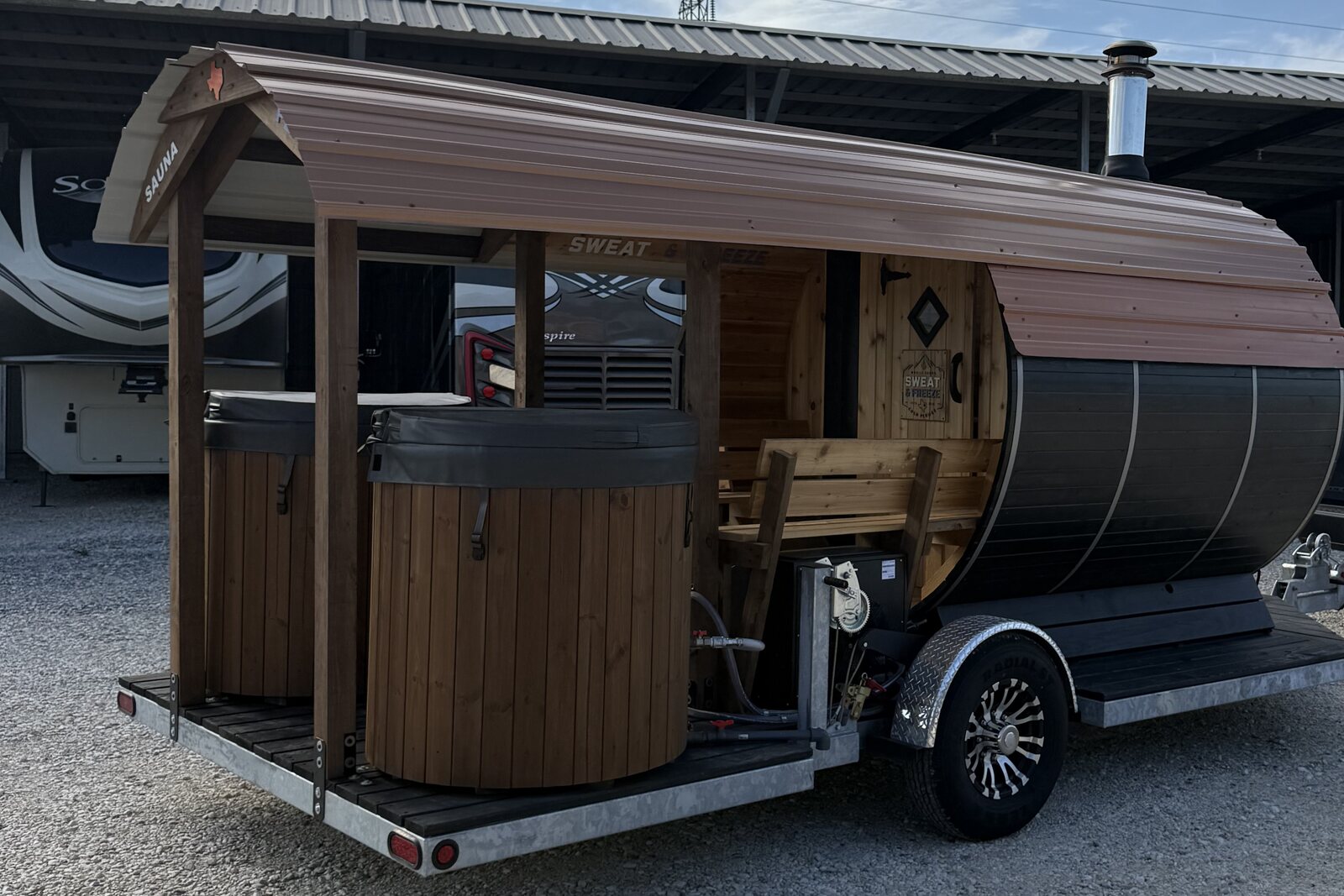 A trailer carrying a barrel sauna with a stovepipe and a covered porch with two cold plunge tubs, parked on gravel in front of a steel shed