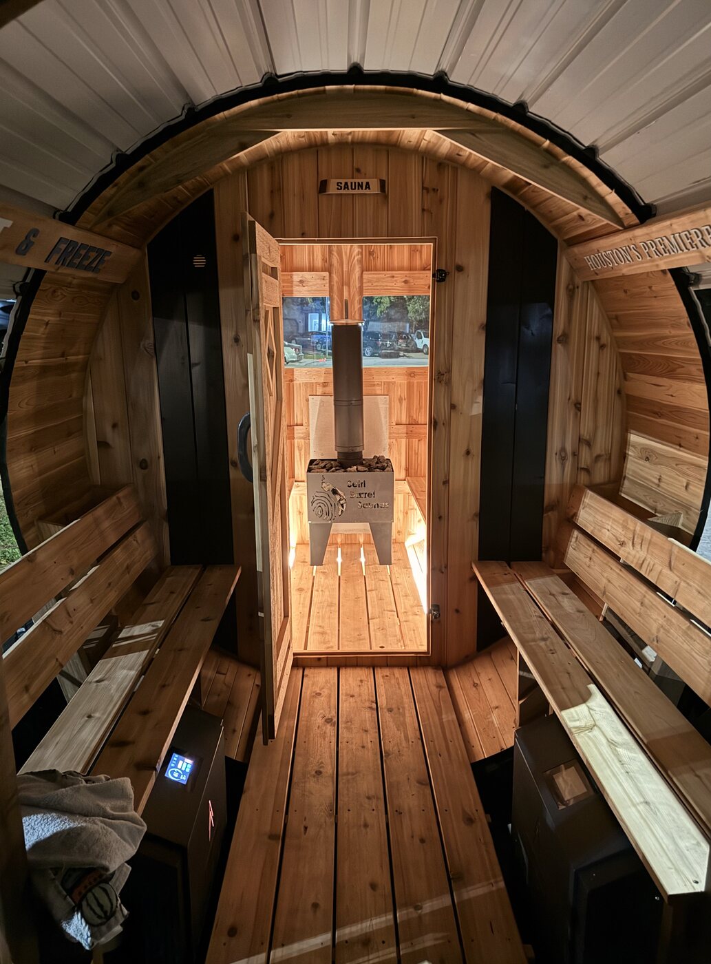 View from a covered porch with benches through an open sauna door to a wood stove at the far end of a barrel sauna