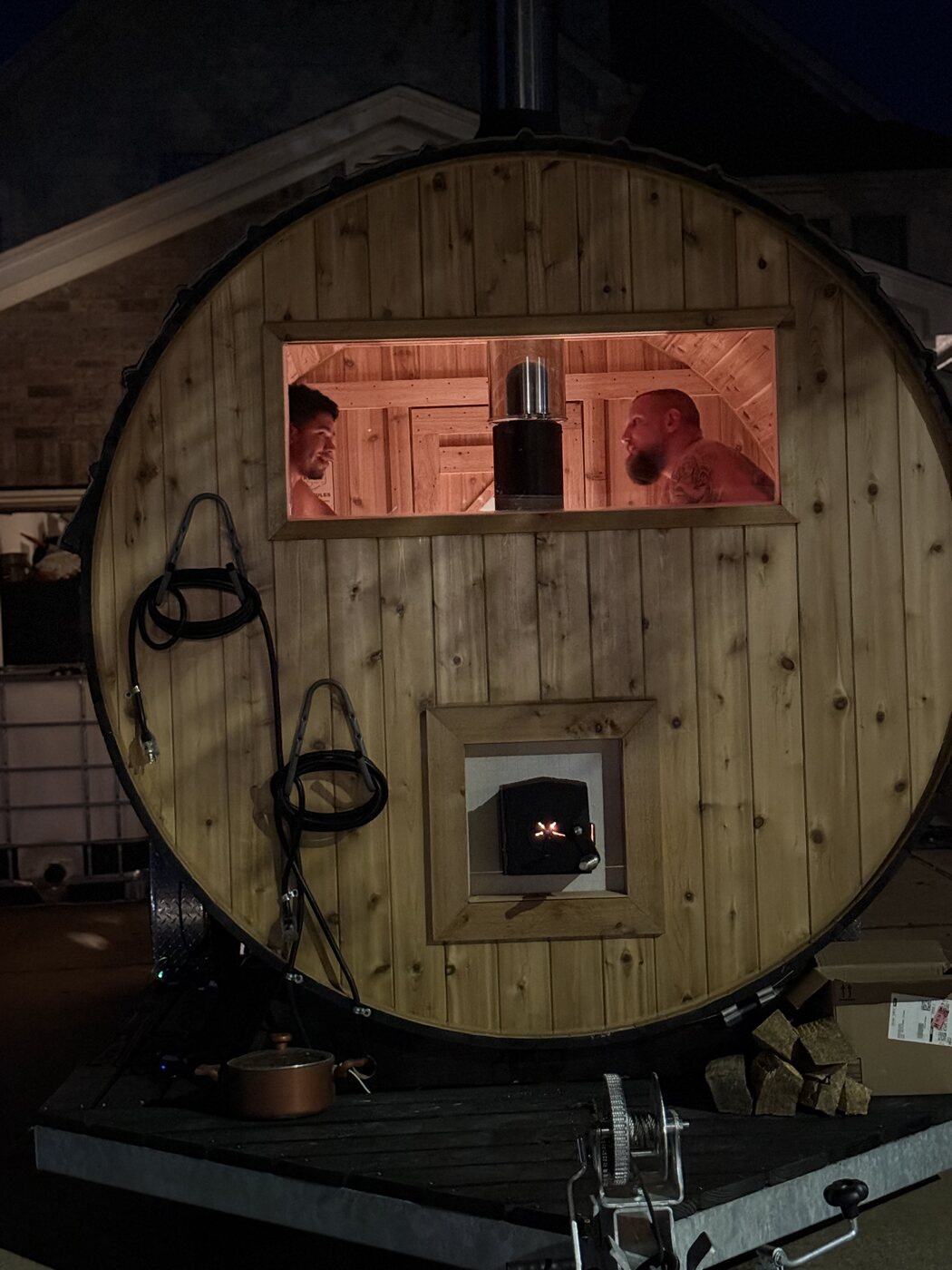 Two bathers talking in the lit window of a barrel sauna at night, with fire glowing in the stove door set into the end wall below