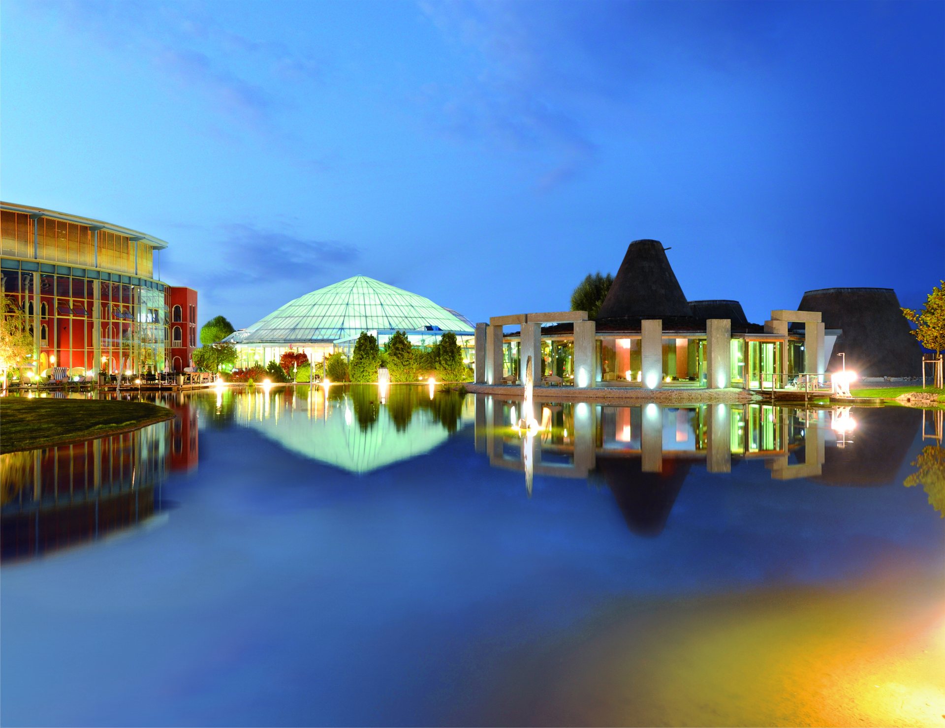 The glass dome of Therme Erding's bathing hall and its sauna garden buildings lit at dusk and reflected in a lake