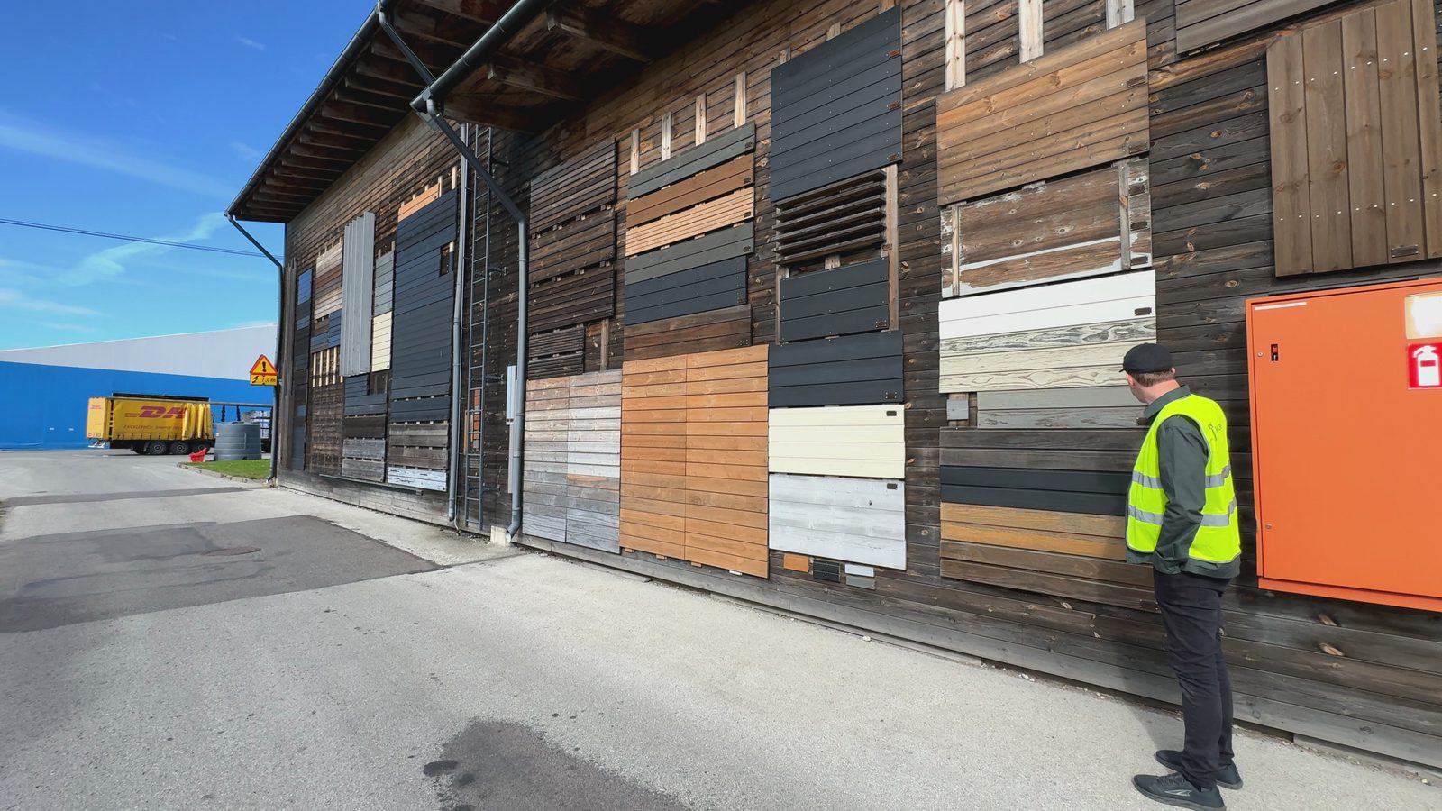 A long timber wall covered in cladding samples in brown, gray, black and white, with a man in a yellow vest studying the boards