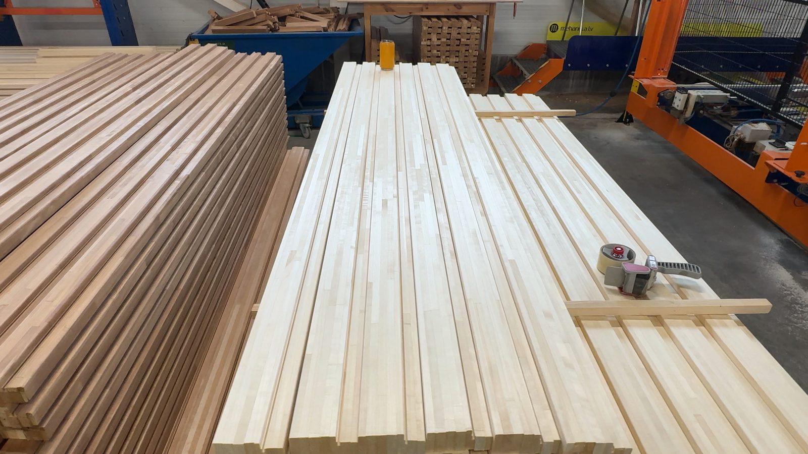 Two long stacks of machined wooden slats on a factory floor, one pale and one darker brown, with an orange machine frame behind