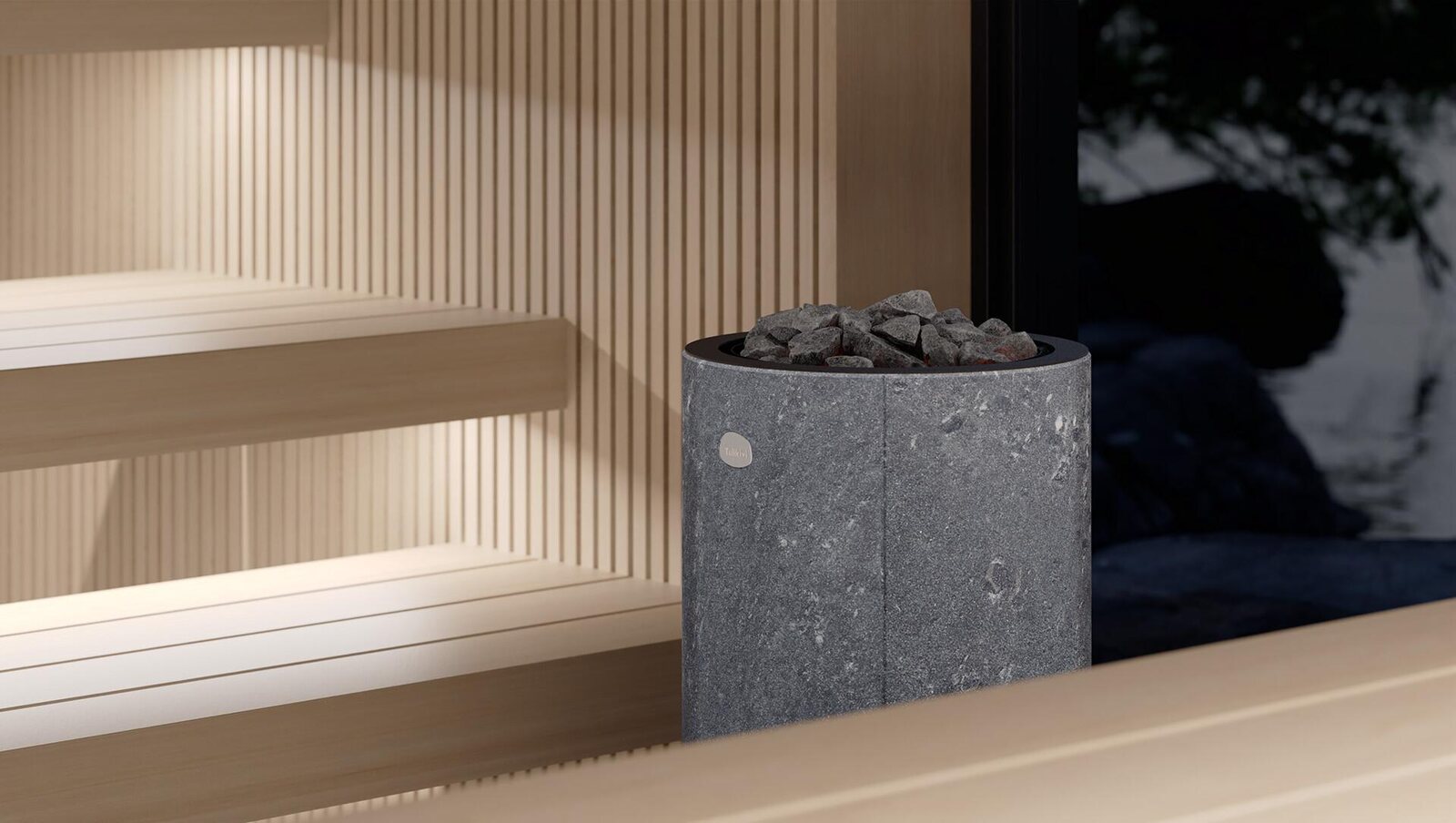 A round grey soapstone Tulikivi Kaarna heater with dark stones on top, beside pale wooden sauna benches and a window