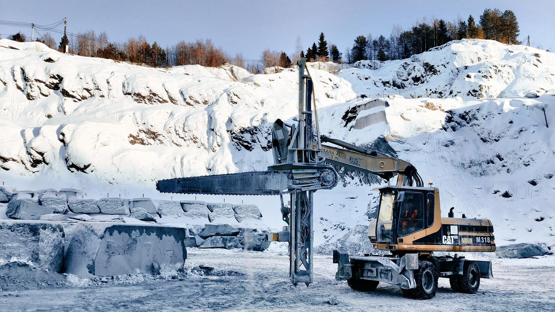 An excavator fitted with a long chain-saw arm cuts soapstone in a snow-covered quarry, with sawn blocks behind it