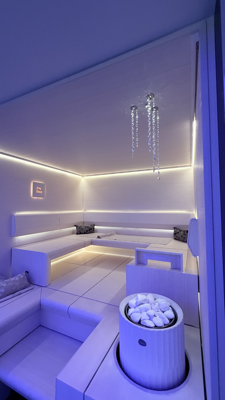 A white cylindrical Tulikivi electric heater filled with white stones beside the benches of a white display sauna lit violet
