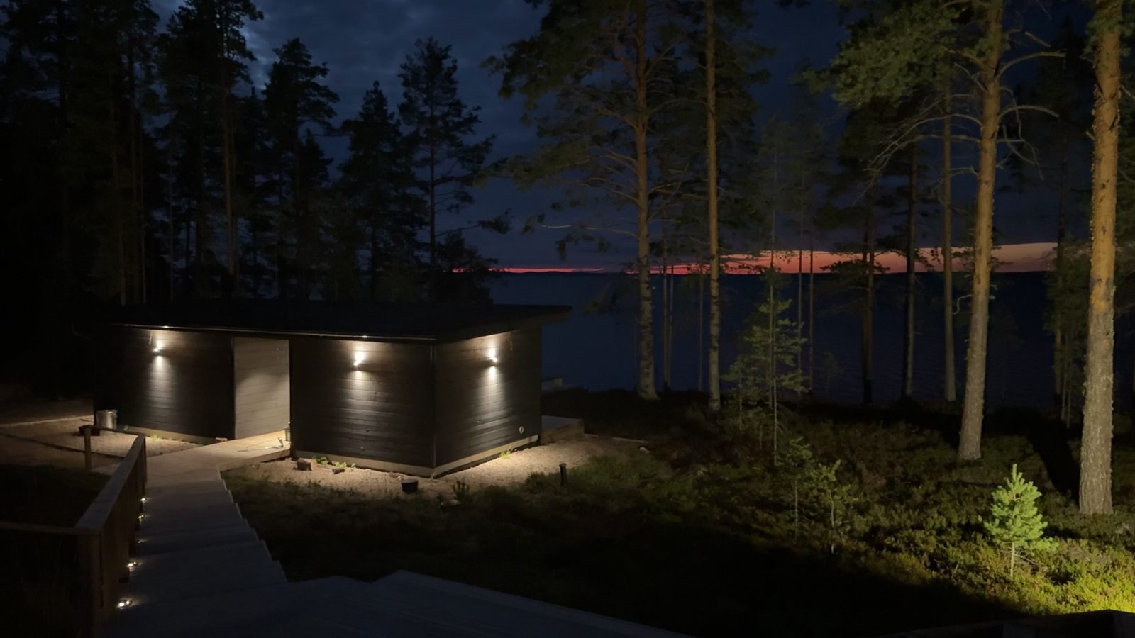 A low black timber sauna building lit by small wall lights among pines at night, with red sky over Lake Saimaa behind it