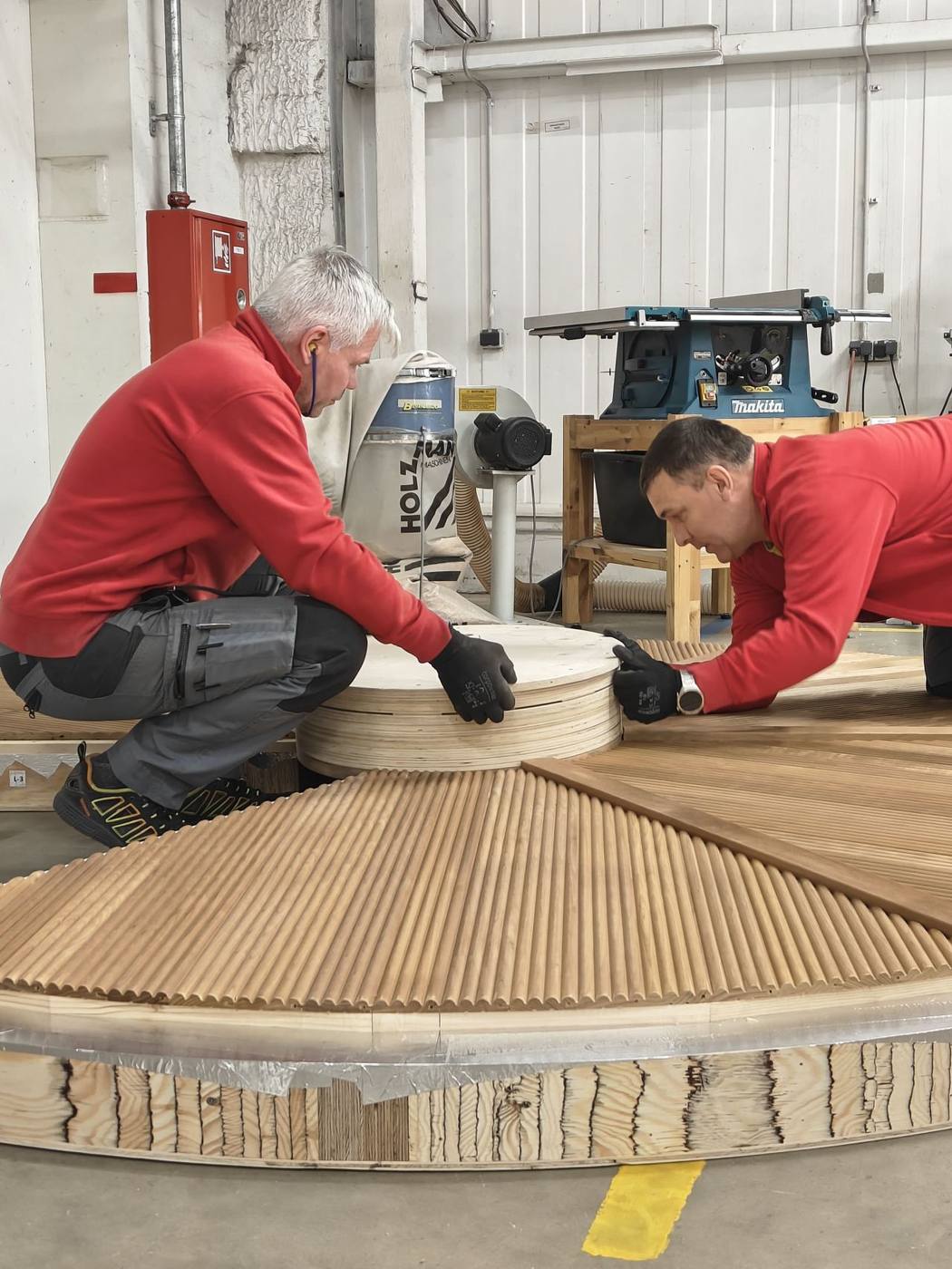 Two Auroom craftsmen kneel beside a curved CNC-machined sauna bench module in the Tartu factory, dust extractor and Makita tools visible behind them