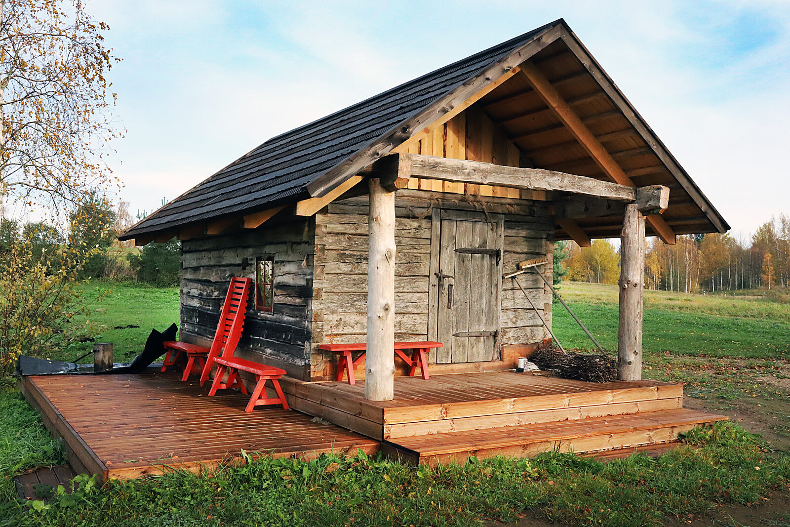A traditional Estonian log smoke sauna with a black-tarred roof and a wooden deck, in autumn