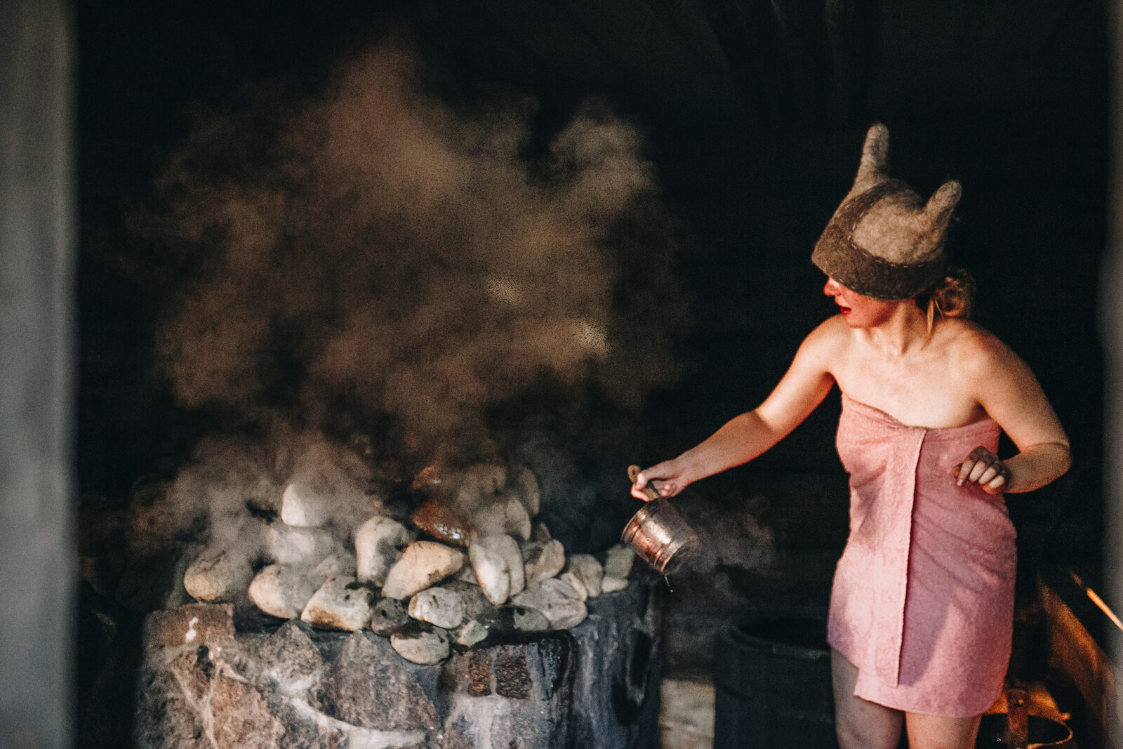 A woman in a felt sauna hat throwing water onto the stones of an Estonian smoke sauna stove